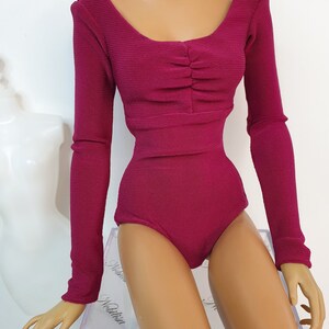 May include: A magenta-colored bodysuit with long sleeves. The scoop neckline is gathered at the bust. The fabric has a ribbed texture. The garment is displayed on a mannequin.