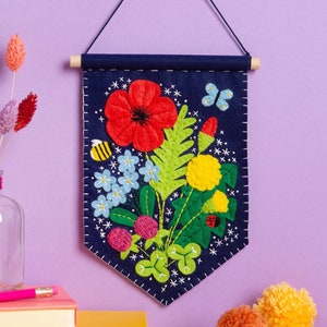 May include: A navy blue felt banner with a floral embroidery design. The banner features a red poppy, yellow flowers, green leaves, and a bee. The banner is hanging on a purple wall.