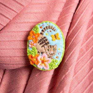 May include: A felt pin with a green, blue and yellow background featuring a brown and white cat with a butterfly and flowers.