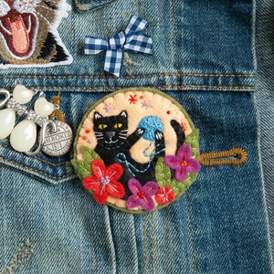 May include: A black felt cat with yellow eyes playing with a blue yarn ball. The cat is surrounded by colourful felt flowers and green leaves. The felt applique is on a denim background.