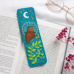 May include: Teal felt bookmark with a brown owl design, a white crescent moon, and embroidered stars. The owl is perched on a green leafy branch with red berries. The bookmark is placed on an open book.