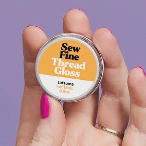 May include: A small, round metal container of Sew Fine Thread Gloss, labeled "satsuma" and "net 15ml 0.5oz." The container has a white and orange label with black text. The background is a solid purple.