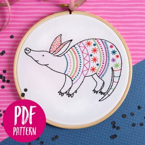 Può includere: Un motivo di ricamo colorato di un armadillo con un corpo bianco e un guscio con un motivo colorato. Il motivo è in un cerchio e il testo "PDF PATTERN" è su uno sfondo rosa.