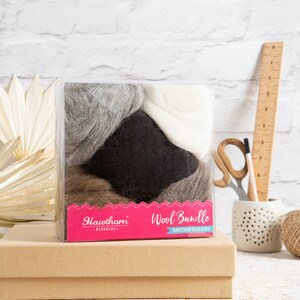 May include: A clear plastic box filled with four different colors of wool: gray, white, brown, and black. The box has a pink label that says "Hawthorn Wool Bundle British Breeds - Handmade".