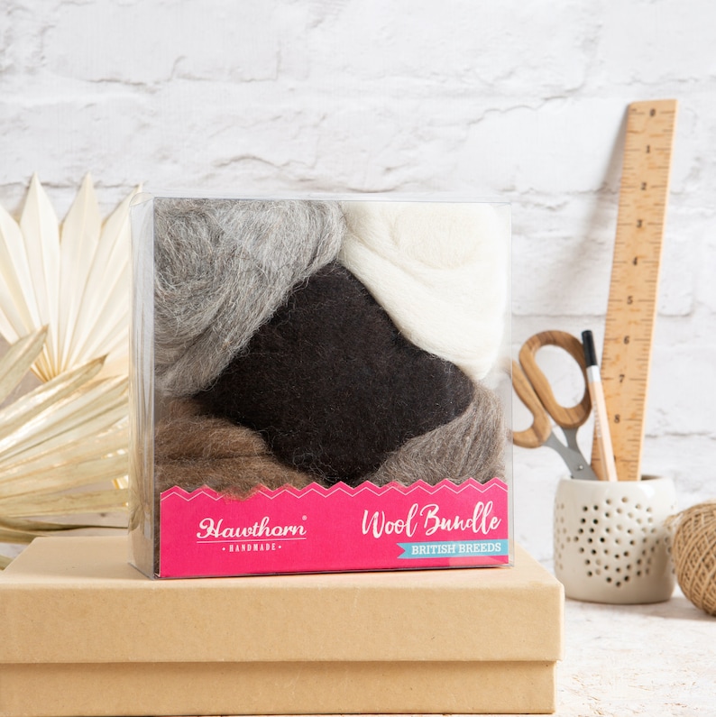 May include: A clear plastic box filled with four different colors of wool: gray, white, brown, and black. The box has a pink label that says "Hawthorn Wool Bundle British Breeds - Handmade".