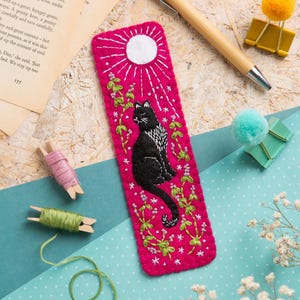 May include: A rectangular fuchsia felt bookmark featuring an embroidered black cat sitting amidst green vines with small purple flowers, a white sun, and white star accents. The edges are finished with a decorative blanket stitch.