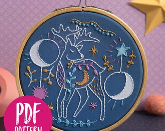 May include: Embroidered hoop art featuring a white deer with floral details on a dark blue background. The deer is surrounded by white crescent moons, stars, and other floral elements. The hoop is made of wood and has a gold metal stand.