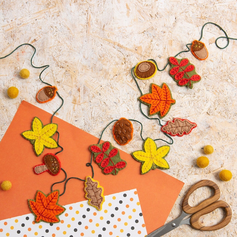 May include: A felt garland with autumn leaves, acorns, and other fall-themed decorations. The garland is strung on a twine cord and is displayed on a light brown wood background.