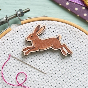 May include: A wooden hare-shaped embellishment in shades of brown, placed on white cross-stitch fabric within an embroidery hoop. A needle and pink thread are also visible, suggesting a crafting project. The hare is in mid-leap.