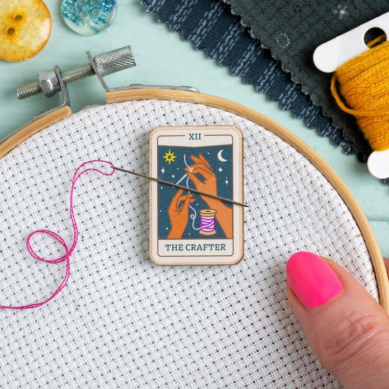 Pu&ograve; includere: Un telaio da ricamo in legno tiene un tessuto bianco con filo rosa e un ago. Una carta dei tarocchi con "THE CRAFTER" mostra mani, una bobina di filo ed elementi celesti. Sono visibili altri materiali per l'artigianato.