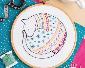 Puede incluir: Un bastidor de bordado con un diseño de gato durmiente. El gato blanco lleva un suéter bordado de colores. El bastidor de madera está rodeado de materiales de costura, incluyendo una cinta métrica e hilo.