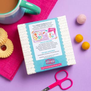 a box of embroidery kit, a pair of scissors, a mug of coffee, cookies, and a pink and white patterned cloth on a purple background.
