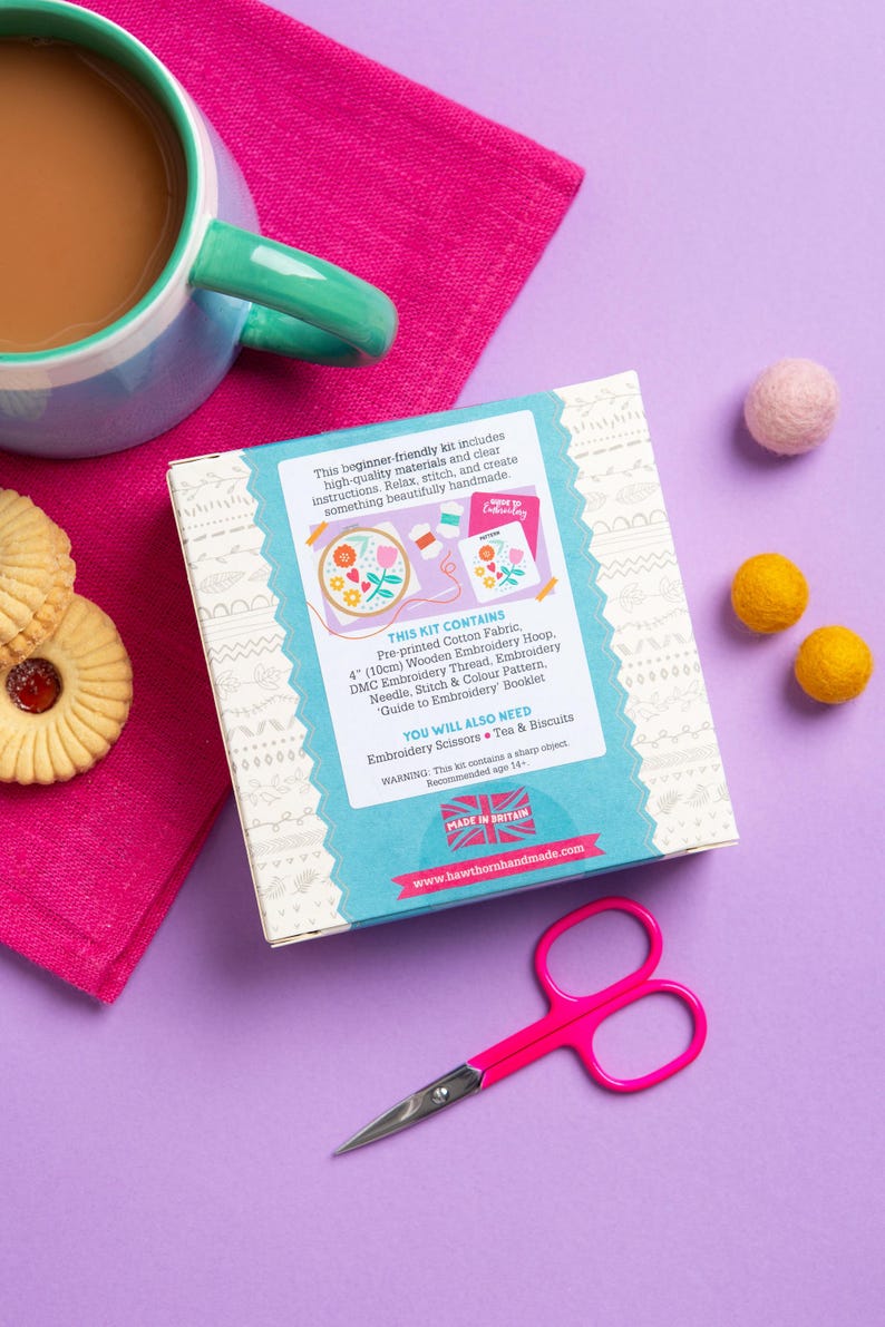 a box of embroidery kit, a pair of scissors, a mug of coffee, cookies, and a pink and white patterned cloth on a purple background.