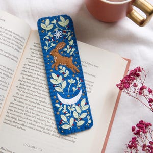 May include: A blue felt bookmark with a brown rabbit, white crescent moon, and floral embroidery. The bookmark is rectangular and features a detailed, handcrafted design with a whimsical theme.