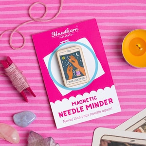 Pu&ograve; includere: Un magnete per aghi rosa con il testo "MAGNETIC NEEDLE MINDER" e "Never lose your needle again!". Il promemoria presenta un disegno di carta dei tarocchi con il testo "THE CRAFTER". Lo sfondo &egrave; rosa con strisce rosa e bianche. Sono visibili anche una bobina di filo e una candela.