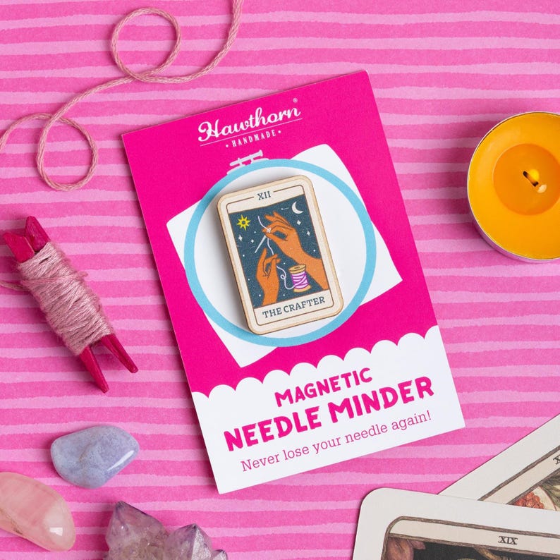 Pu&ograve; includere: Un magnete per aghi rosa con il testo "MAGNETIC NEEDLE MINDER" e "Never lose your needle again!". Il promemoria presenta un disegno di carta dei tarocchi con il testo "THE CRAFTER". Lo sfondo &egrave; rosa con strisce rosa e bianche. Sono visibili anche una bobina di filo e una candela.