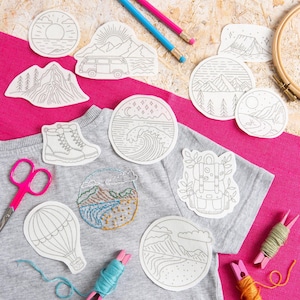 May include: A selection of iron-on patches featuring outdoor scenes, such as mountains, a camper van, and a hot air balloon. The patches are arranged on a grey t-shirt, alongside embroidery tools and pencils.