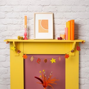 May include: A yellow painted fireplace mantel with a fall-themed garland of felt leaves and acorns. The garland is strung across the top of the mantel. A framed picture with a peach-colored background hangs above the mantel. A pink candle holder with a white candle sits on the mantel.