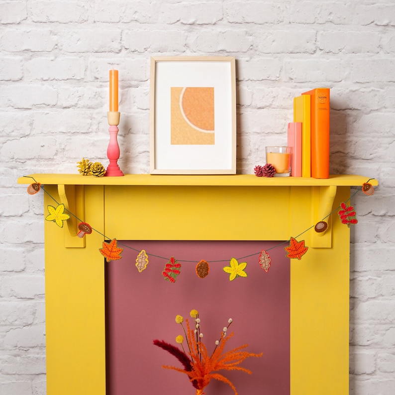 May include: A yellow painted fireplace mantel with a fall-themed garland of felt leaves and acorns. The garland is strung across the top of the mantel. A framed picture with a peach-colored background hangs above the mantel. A pink candle holder with a white candle sits on the mantel.