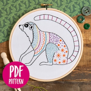 May include: Embroidered hoop art featuring a colourful lemur with a pink tail, sitting on a white background. The lemur is outlined in black and filled with various colourful embroidery stitches.