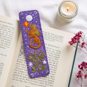 May include: A handmade purple felt bookmark with an embroidered orange dragon breathing fire. The bookmark features a white moon, stars, and floral details. A lit candle and open book are in the background.