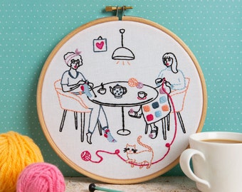 Puede incluir: Arte de bordado en aro que representa a dos personas sentadas en una mesa con un gato, tazas de té e hilo. La escena está bordada en hilo negro sobre tela blanca con acentos de colores.