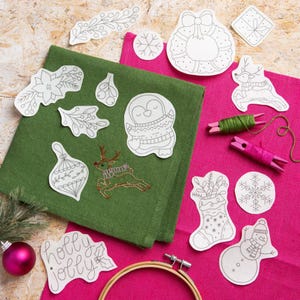 May include: A selection of Christmas embroidery designs and materials. Features a green cloth with an embroidered reindeer, a pink cloth, and paper cutouts of festive ornaments, a penguin, and a snowman. Also includes embroidery thread and a wooden hoop.