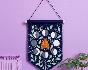 May include: A dark blue felt wall hanging featuring an embroidered orange moth at the center, surrounded by light green leaves, white star accents, and various moon phases in shades of gray and white, suspended from a wooden dowel with a string.