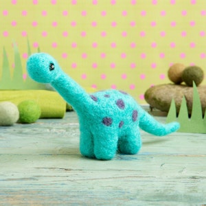 May include: A teal blue felt dinosaur toy with dark purple spots. The dinosaur is standing on a light blue wooden surface.