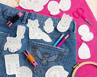 May include: A denim jean pocket with a detailed embroidered owl design, surrounded by various craft supplies. Includes paper cutouts of animals and floral designs, pencils, scissors, embroidery floss, and a wooden embroidery hoop. The background is a bright pink fabric.