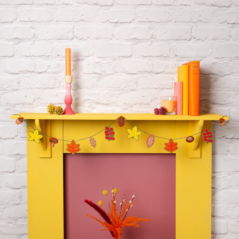 May include: A yellow painted fireplace mantel with a garland of crocheted fall leaves and mushrooms. The garland is strung with a brown twine. The fireplace has a pink painted interior.