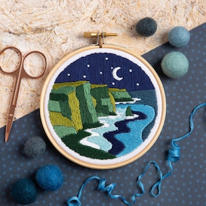 以下が含まれることがあります： 木製のフレームに刺繍された海岸の風景。夜空の下の崖、三日月と星が描かれています。青、緑、白の色使い。銅製のハサミと、さまざまな青色のフェルトボールも写っています。