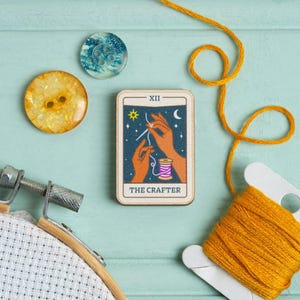 Pu&ograve; includere: Un'immagine a tema artigianale con una carta dei tarocchi in legno con la scritta "THE CRAFTER", due bottoni, una bobina di filo giallo e un telaio per punto croce. La carta dei tarocchi raffigura mani che tengono ago e filo.