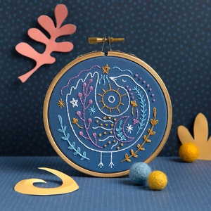May include: A blue embroidery hoop with a white, pink, and yellow embroidered design of a bird with a sun and moon inside. The design is surrounded by small flowers and leaves.
