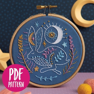 Puede incluir: Un aro de bordado azul con un diseño de conejo blanco, rodeado de estrellas, lunas y elementos florales en hilo dorado, rosa y azul. El aro está sobre un fondo morado y azul.
