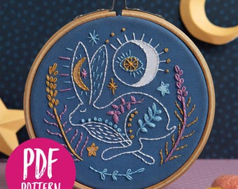 May include: A blue embroidery hoop with a white rabbit design, surrounded by stars, moons, and floral elements in gold, pink, and blue thread. The hoop is on a purple and blue background.