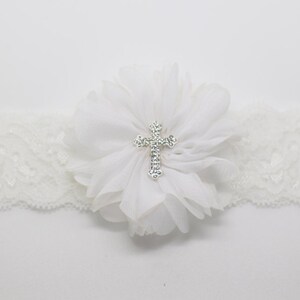 May include: Ivory lace garter featuring a large white fabric flower with a sparkling silver cross in the center. The garter is designed with a scalloped edge and is suitable for a special occasion.