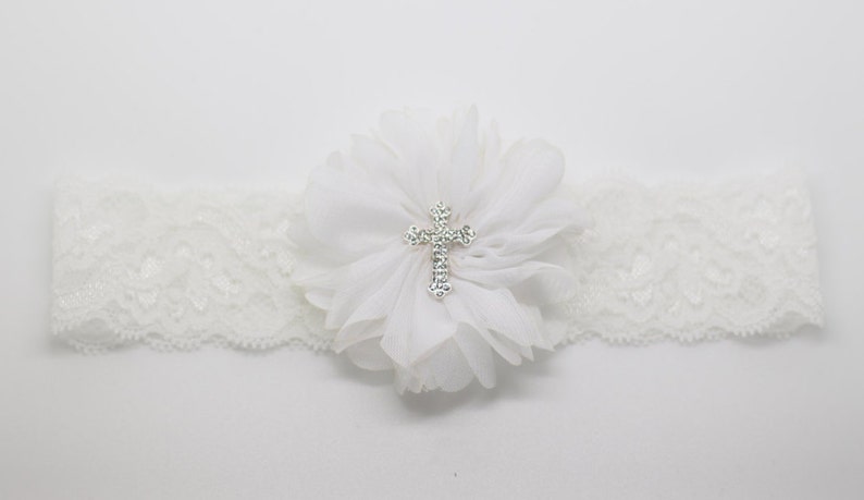 May include: Ivory lace garter featuring a large white fabric flower with a sparkling silver cross in the center. The garter is designed with a scalloped edge and is suitable for a special occasion.