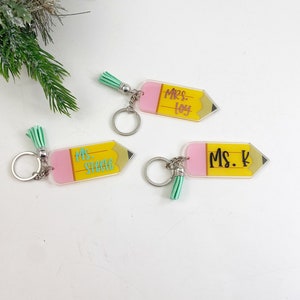 Puede incluir: Tres llaveros en forma de l&aacute;piz amarillo con detalles en rosa y verde. Los llaveros tienen los nombres "Mrs. Loy", "Ms. Steele" y "Ms. K" escritos en ellos.