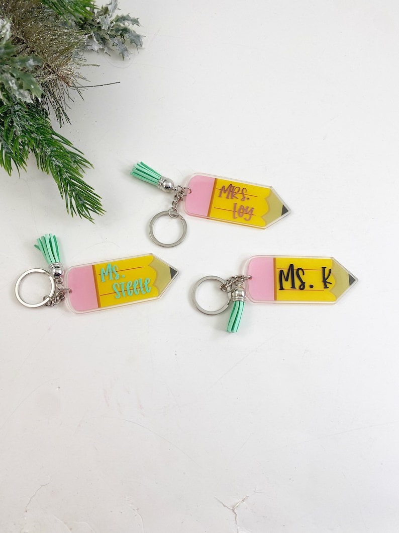 Puede incluir: Tres llaveros en forma de l&aacute;piz amarillo con detalles en rosa y verde. Los llaveros tienen los nombres "Mrs. Loy", "Ms. Steele" y "Ms. K" escritos en ellos.