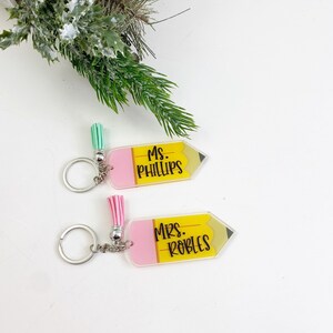 Puede incluir: Dos llaveros en forma de l&aacute;piz amarillo con gomas de borrar rosas. El llavero superior dice "Ms. Phillips" y el llavero inferior dice "Mrs. Robles".