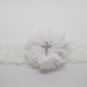 May include: Ivory lace garter with a white chiffon flower and a silver cross embellished with rhinestones. The garter is designed for a special occasion, such as a wedding or baptism.