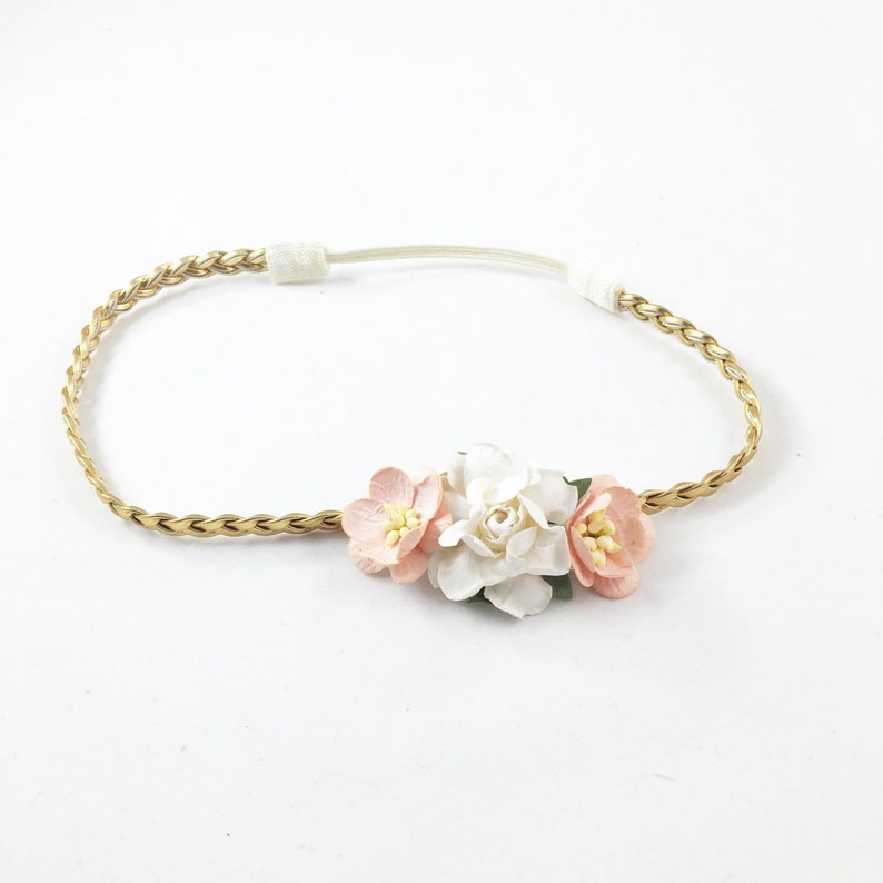 May include: A gold-colored braided headband with a cluster of fabric flowers. The flowers are in shades of white and peach, with yellow centers. The headband is on a white background.