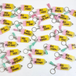 Puede incluir: Una colecci&oacute;n de llaveros en forma de l&aacute;piz amarillo y rosa con la palabra "Miss" seguida de un nombre en cada llavero. Los llaveros tienen un borla rosa atada a cada uno.