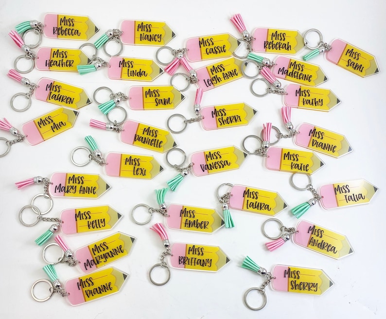 Puede incluir: Una colecci&oacute;n de llaveros en forma de l&aacute;piz amarillo y rosa con la palabra "Miss" seguida de un nombre en cada llavero. Los llaveros tienen un borla rosa atada a cada uno.