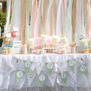 May include: A white table with a white tablecloth and a green and gold banner that says "Happy Birthday". The table is decorated with a variety of treats, including candy, cupcakes, and cotton candy.