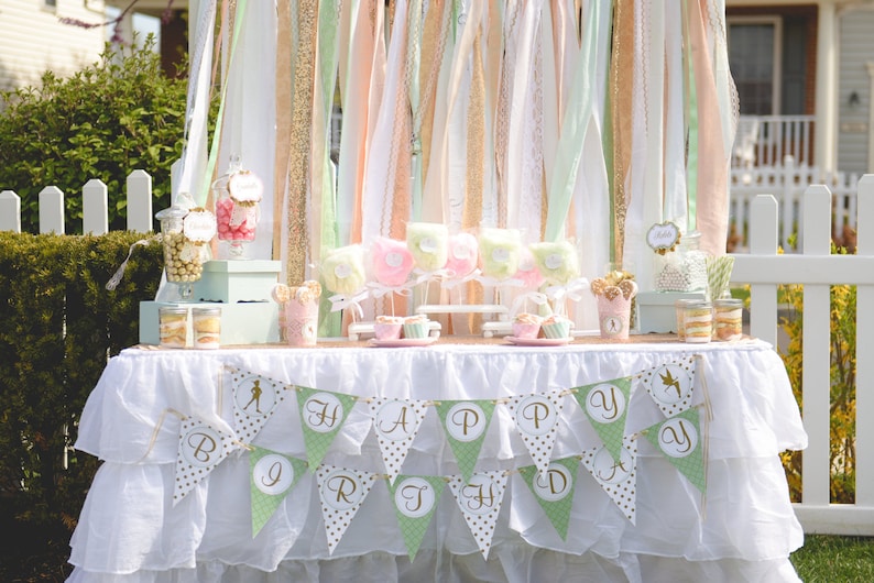 May include: A white table with a white tablecloth and a green and gold banner that says "Happy Birthday". The table is decorated with a variety of treats, including candy, cupcakes, and cotton candy.