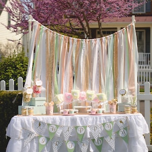 May include: A white table with a white ruffled tablecloth and a green and gold banner that says "Happy Birthday". The table is decorated with pink and green candy, jars of candy, and a white backdrop of fabric streamers.