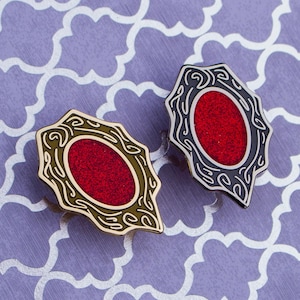 Op de afbeelding: Twee sierlijke emaille pins met een rode kern en decoratieve rand. Eén pin heeft een goudkleurige rand, de andere een zwarte rand. De pins zijn geplaatst tegen een paarse achtergrond met patronen.