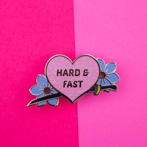 May include: Pink heart-shaped enamel pin with the text "HARD & FAST" in black lettering. The pin is surrounded by two blue flowers and a silver sword.